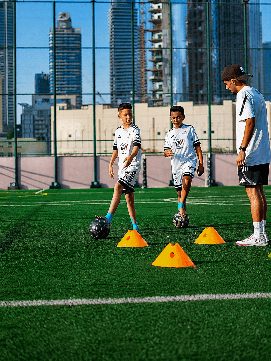 Two players dribbling through orange cones as a coach watches from the touchline
